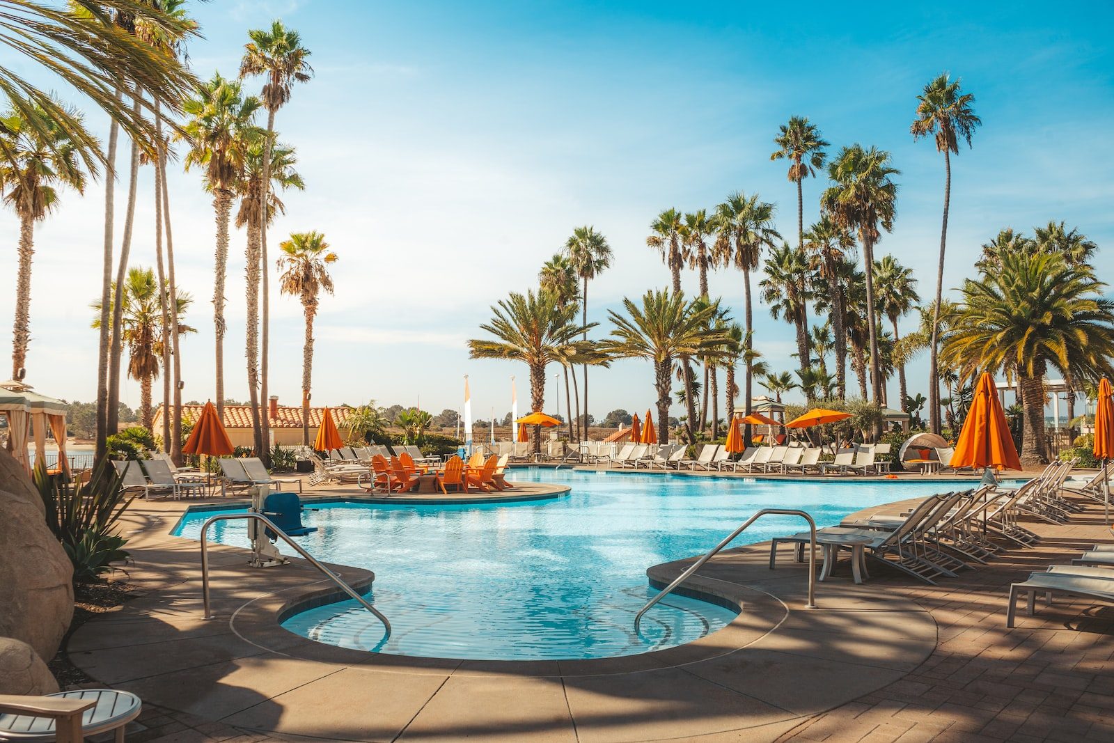 Outdoor resort swimming pool with sun loungers and orange parasols among tall palm trees under a clear blue sky