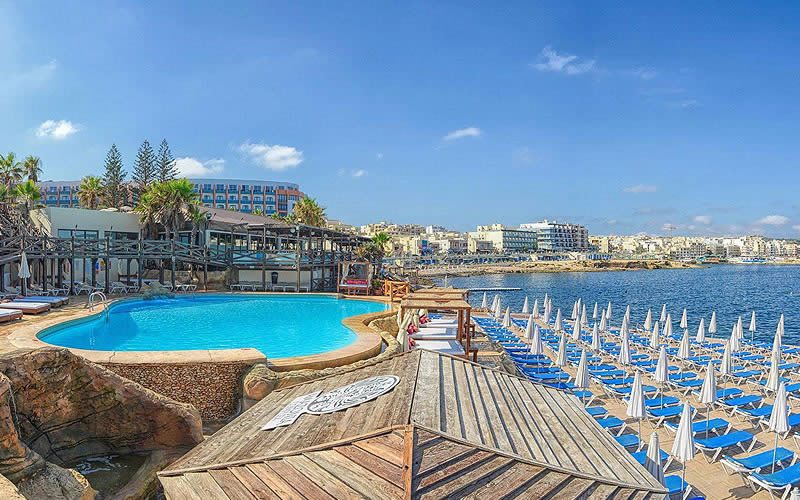 Seaside resort in Malta with a curved swimming pool, wooden sun deck and rows of blue loungers and white parasols beside the sea under a clear sky