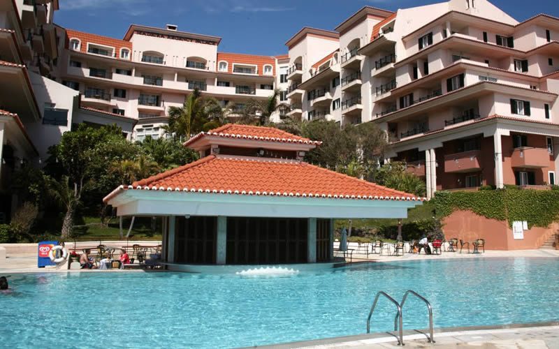 Outdoor swimming pool with a tiled roof pavilion and the Madeira Regency Palace hotel building in the background, Portugal