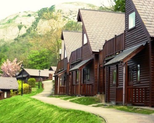 Row of wooden lodge cabins beside a paved path on a grassy hillside with trees and mountains in the background