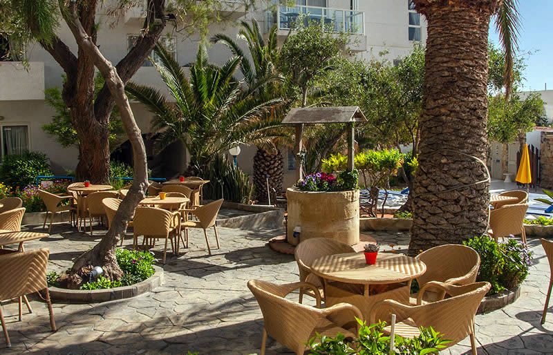 Outdoor courtyard seating area with wicker tables and chairs, palm trees, a stone well planter and a swimming pool beside an apartment building