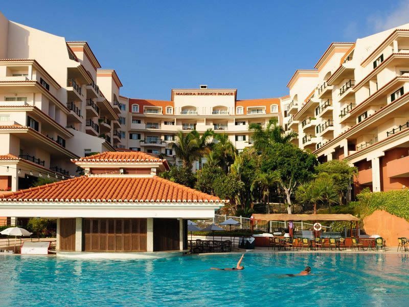 Outdoor swimming pool with loungers in front of the Madeira Regency Palace hotel building under a clear blue sky