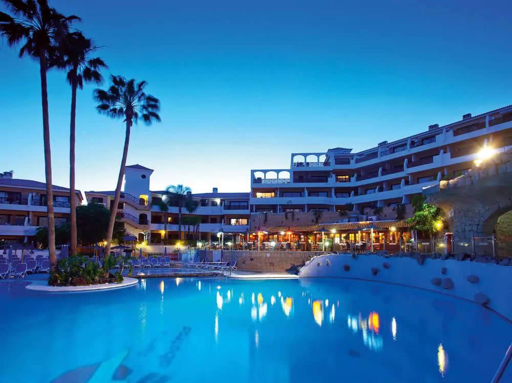 Evening view of a resort hotel with palm trees and a lit swimming pool in the foreground under a clear blue sky