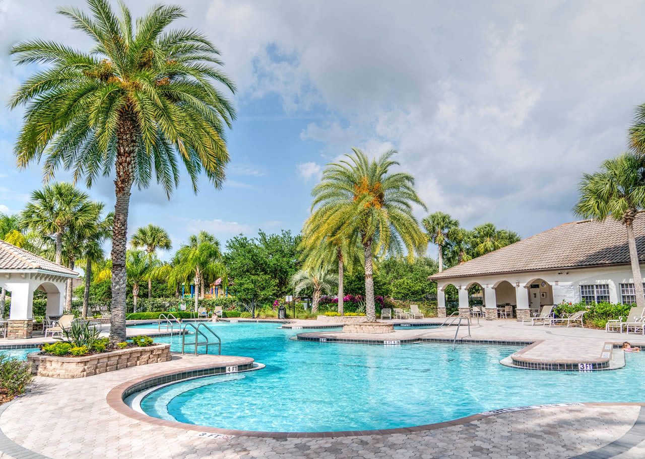 Swimming pool at a holiday resort with palm trees, sun loungers and low white buildings under a partly cloudy sky