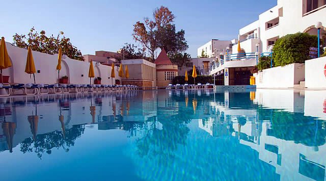Outdoor swimming pool with sun loungers and closed parasols beside white resort buildings under a clear blue sky