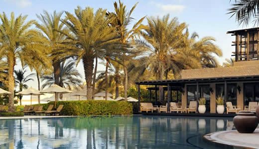 Resort swimming pool with sun loungers and palm trees beside a low modern building in warm evening light