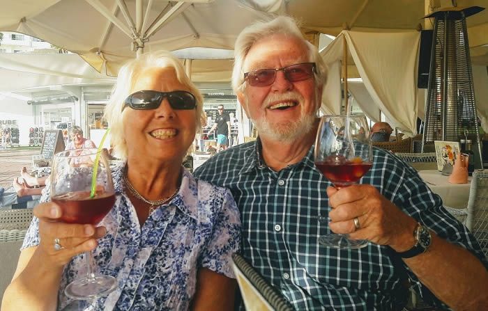 Smiling older couple wearing sunglasses raise glasses of red wine while seated under parasols at an outdoor café terrace