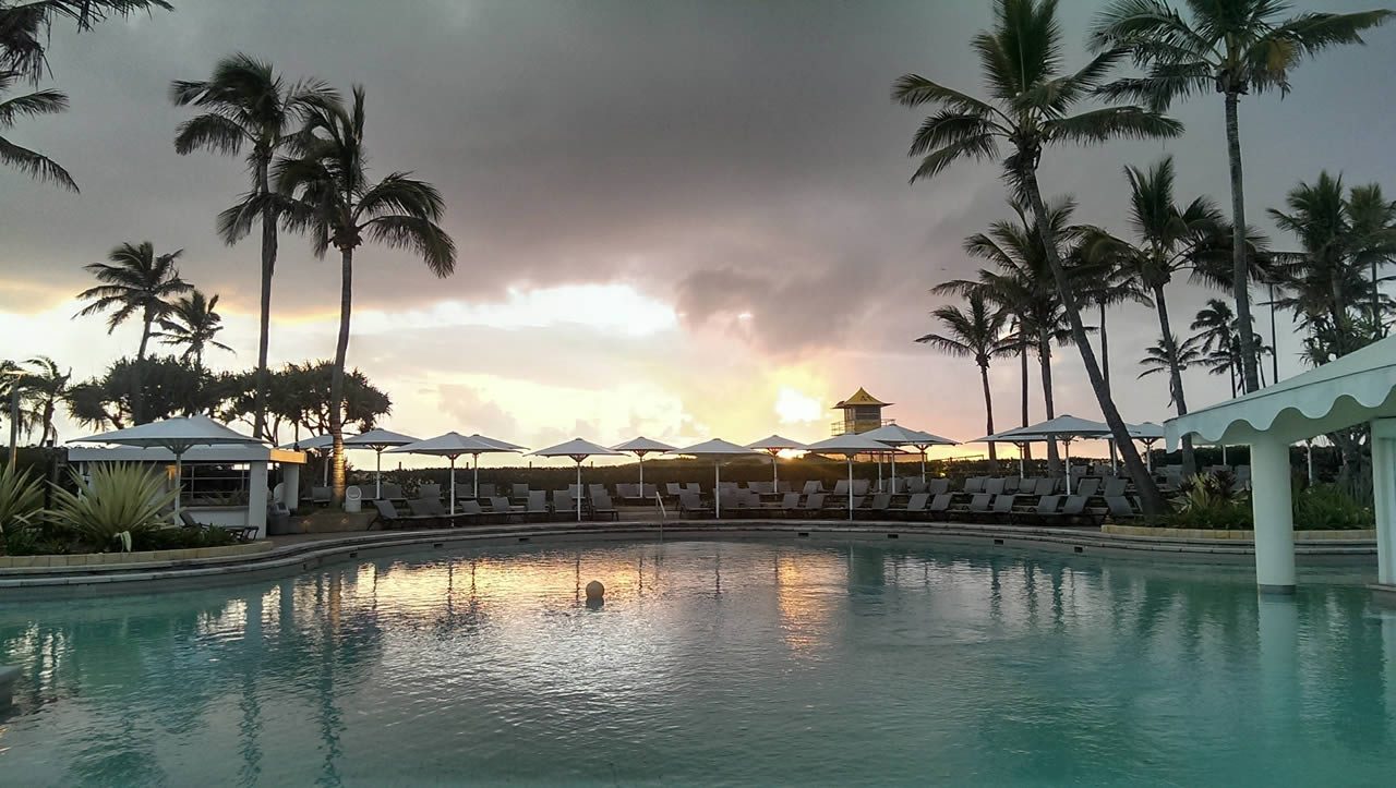 Sunset over a resort swimming pool with palm trees, sun loungers and parasols under dark clouds