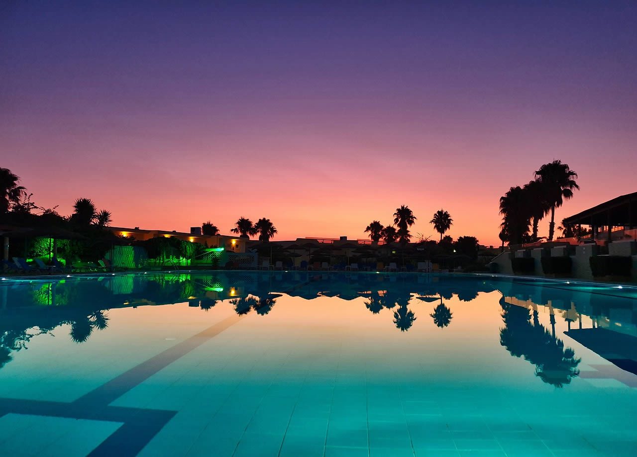 Swimming pool at dusk with palm tree silhouettes and resort buildings reflected in calm water under a purple and orange sunset sky