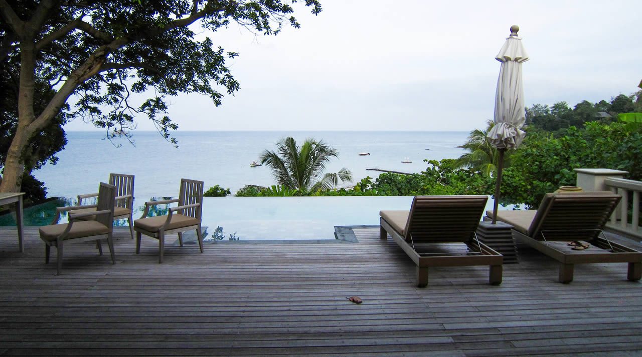 Poolside wooden deck with sun loungers and chairs overlooking the sea, framed by trees and greenery