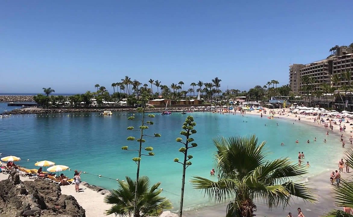 Crowded beach and turquoise lagoon with sun loungers, palm trees and a large resort building in the background under a clear blue sky