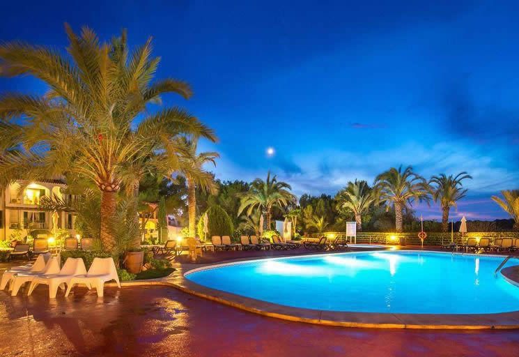Illuminated resort swimming pool at dusk with palm trees, sun loungers and a deep blue sky in Cala Pi, Majorca