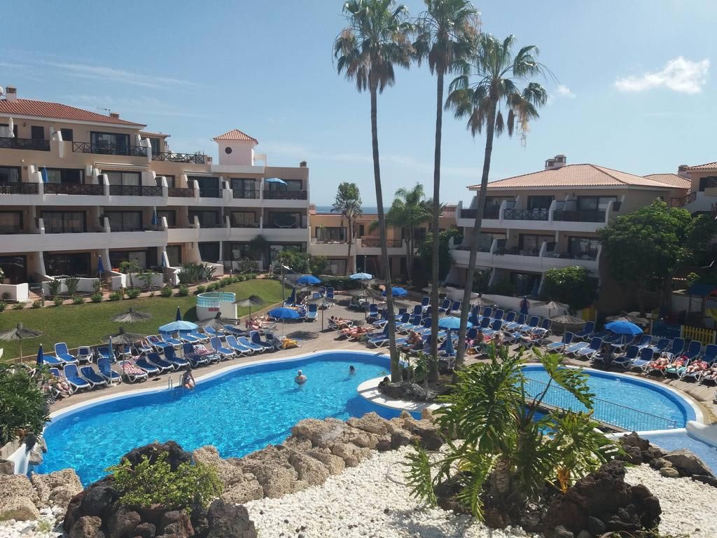 Outdoor hotel pool surrounded by sun loungers, parasols and palm trees between multi-storey apartment buildings on a sunny day
