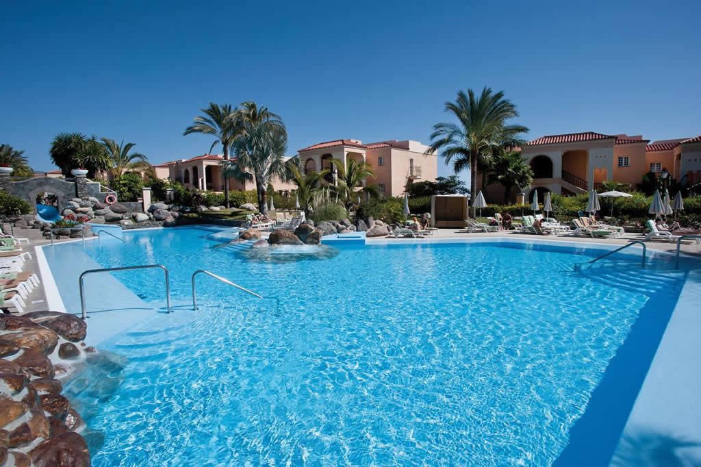 Outdoor resort swimming pool with sun loungers, parasols, palm trees and terracotta-roofed buildings under a clear blue sky