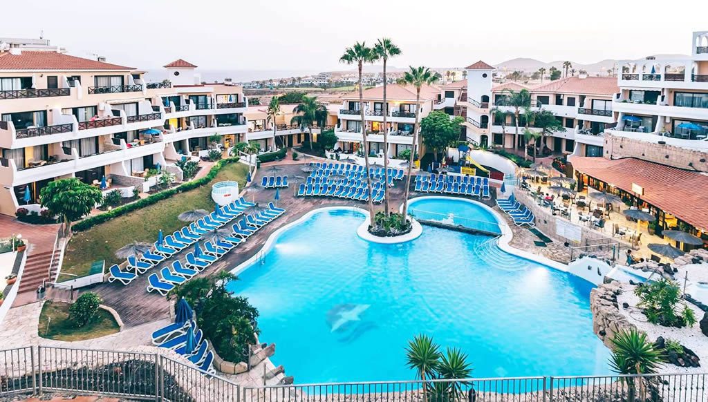 Aerial view of the Parque Albatros resort with a large swimming pool, rows of blue sun loungers, palm trees and white apartment buildings with terracotta roofs.