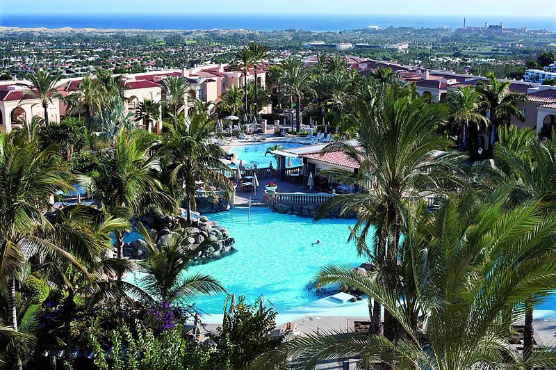 Palm-lined resort swimming pool with sun loungers and terracotta-roofed buildings, overlooking Maspalomas and the sea in Gran Canaria