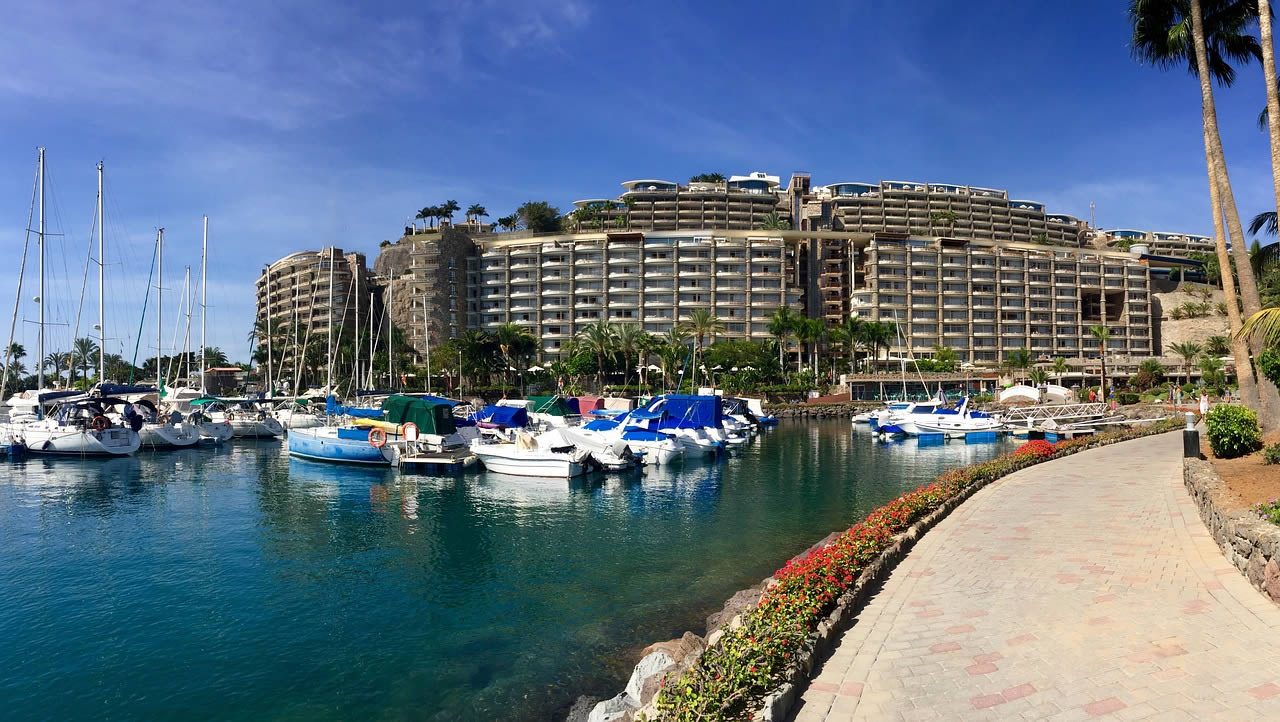 Marina with moored yachts and a waterfront promenade in front of a large resort hotel under a clear blue sky in Gran Canaria