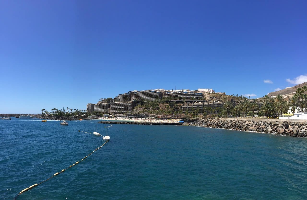 Coastal view in Gran Canaria with blue sea, beach, palm trees and large resort buildings on the hillside under a clear sky