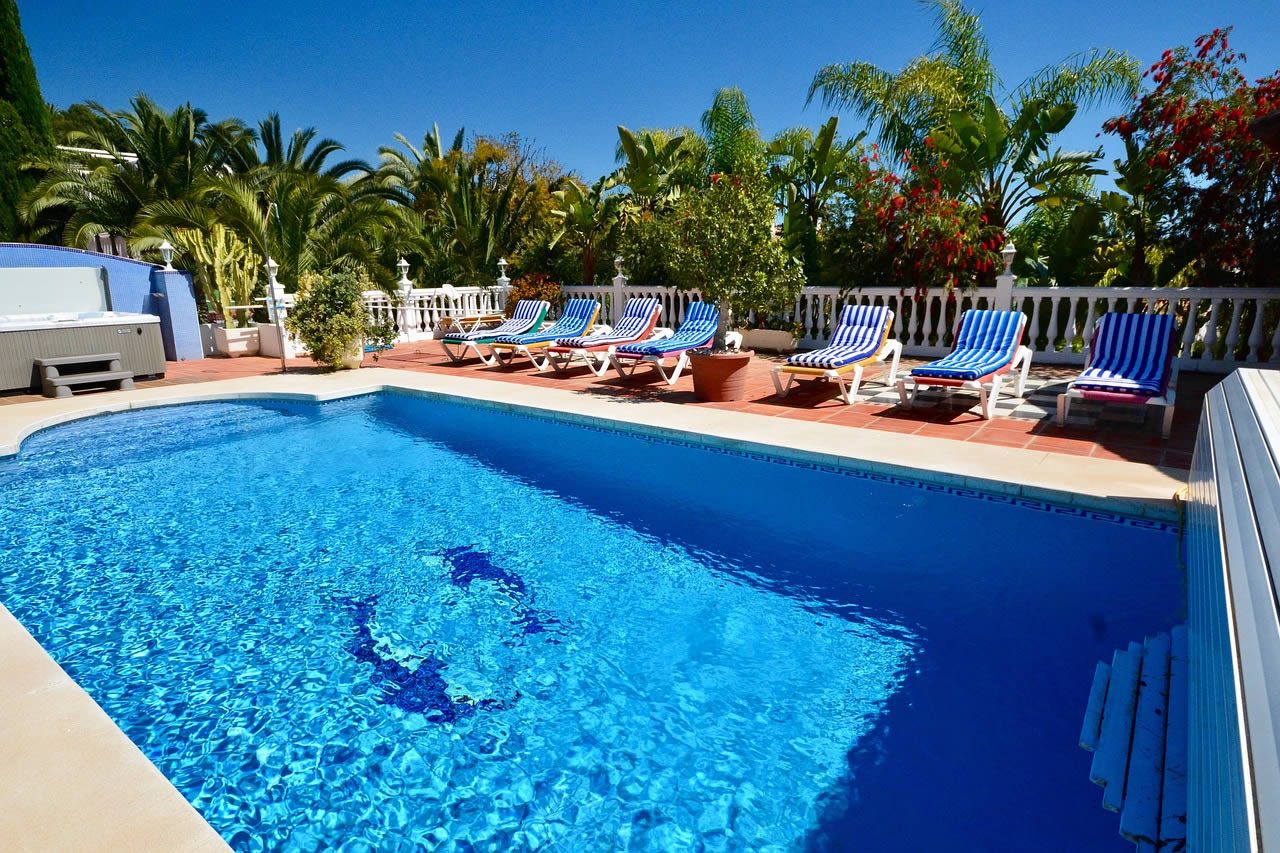 Outdoor resort swimming pool with blue water, striped sun loungers and tropical palm trees under a clear sky