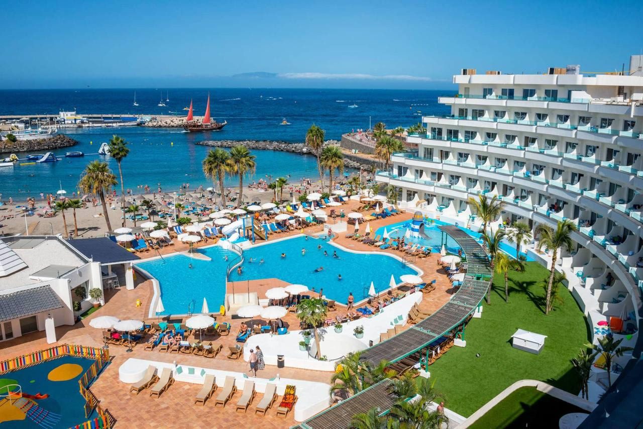 Aerial view of a seaside resort with a large swimming pool, sun loungers and a white multi-storey hotel beside a beach and marina in Tenerife