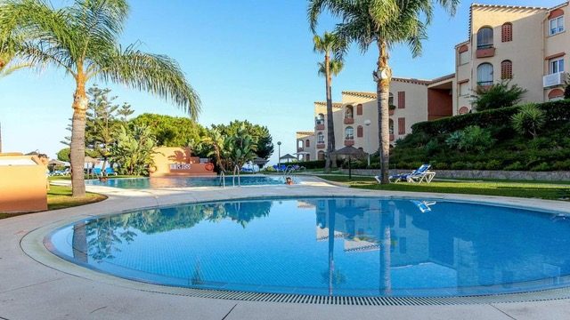Outdoor swimming pool with palm trees, sun loungers and a Spanish-style resort apartment building under a clear blue sky