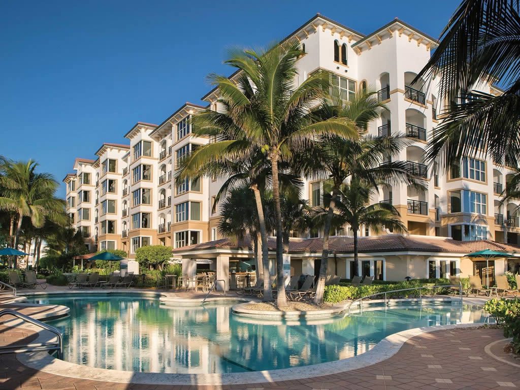 Outdoor swimming pool with sun loungers and palm trees in front of a multi-storey resort building under a clear blue sky
