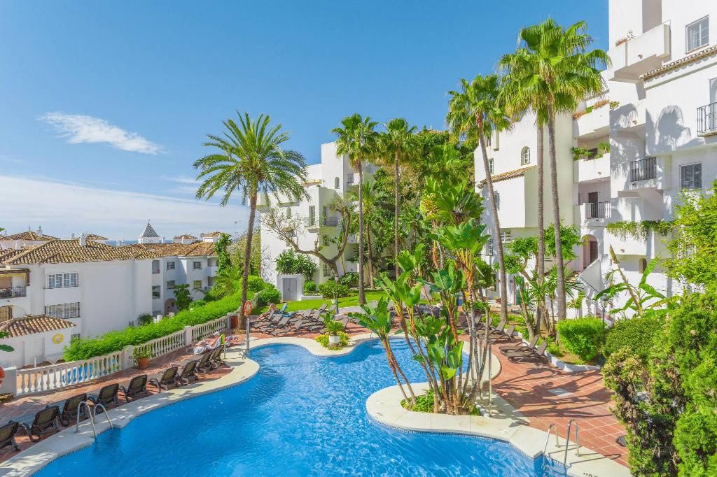 Outdoor resort swimming pool with sun loungers, palm trees and white apartment buildings under a clear blue sky in Spain