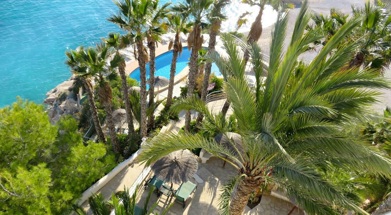 Overhead view of a seaside resort pool area with palm trees, sun loungers and a thatched parasol beside turquoise water