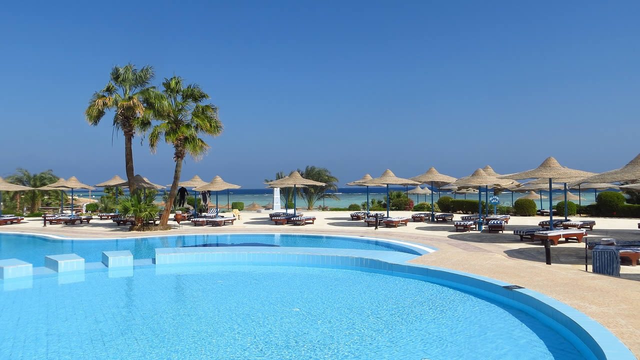 Outdoor resort swimming pool with sun loungers, straw parasols and palm trees overlooking the sea under a clear blue sky