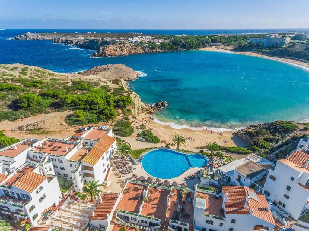 Aerial view of a Spanish coastal resort with terracotta-roof buildings, a large swimming pool and a sandy bay with turquoise sea and rocky headland