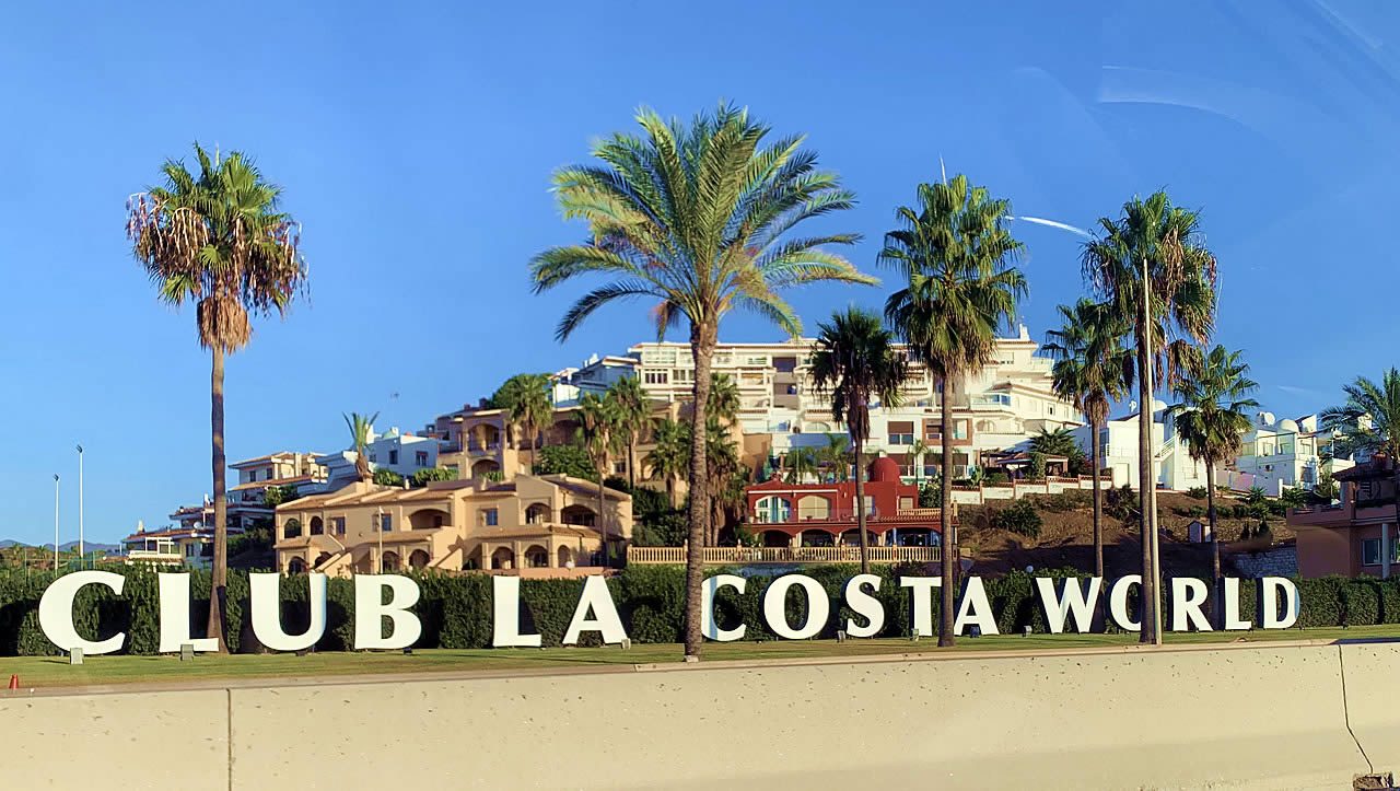 Club La Costa World sign in large white letters in front of palm trees and resort buildings under a clear blue sky