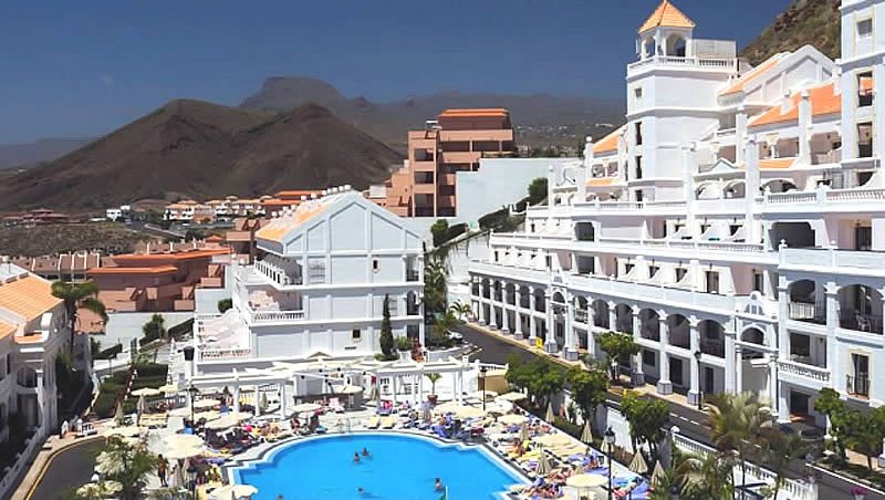 Large hotel complex with balconies beside an outdoor swimming pool and sun loungers, with mountains in the background in Spain