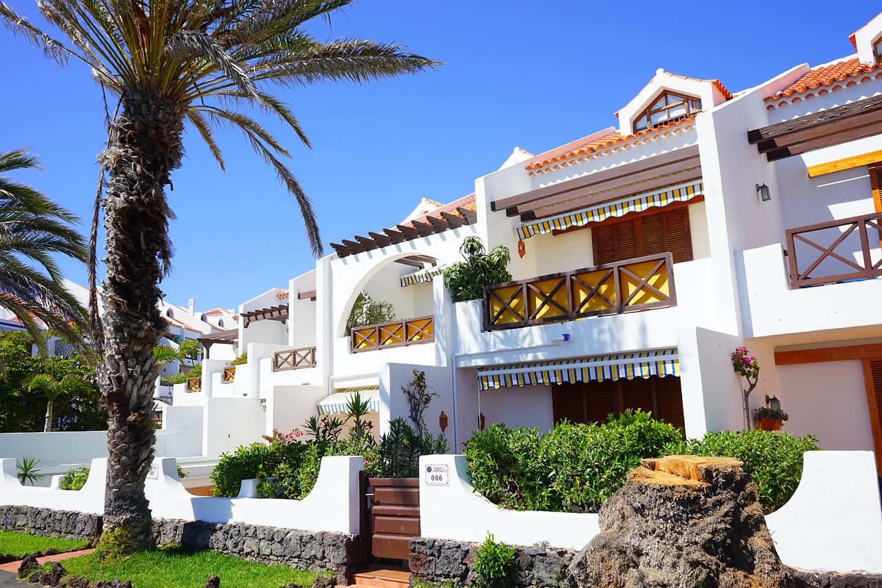 White Mediterranean-style resort apartments with striped awnings and balconies beside a palm tree under a clear blue sky