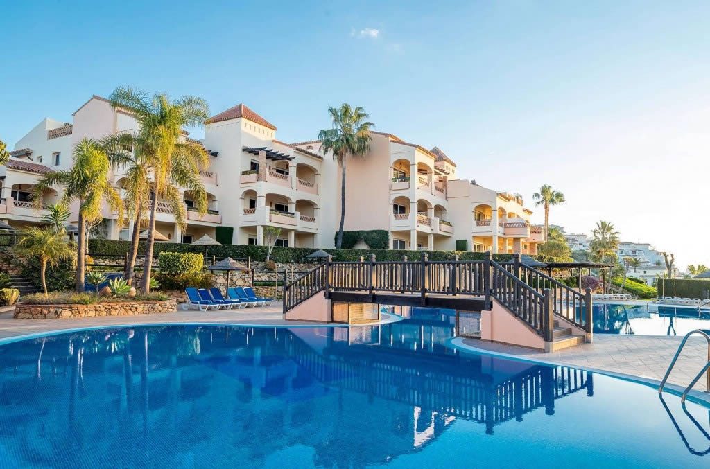 Outdoor swimming pool with a small wooden bridge, sun loungers and palm trees in front of a white resort building under a clear blue sky