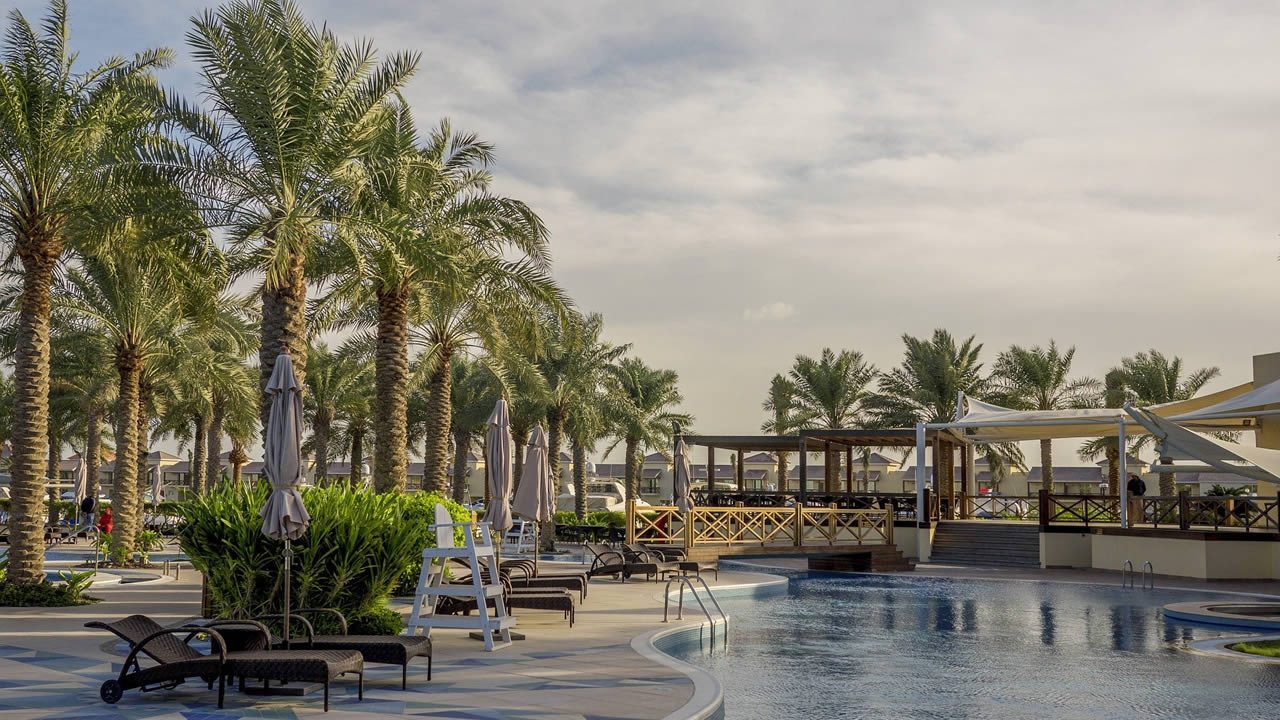Palm-lined resort pool area with sun loungers, closed parasols and a wooden terrace under a cloudy sky