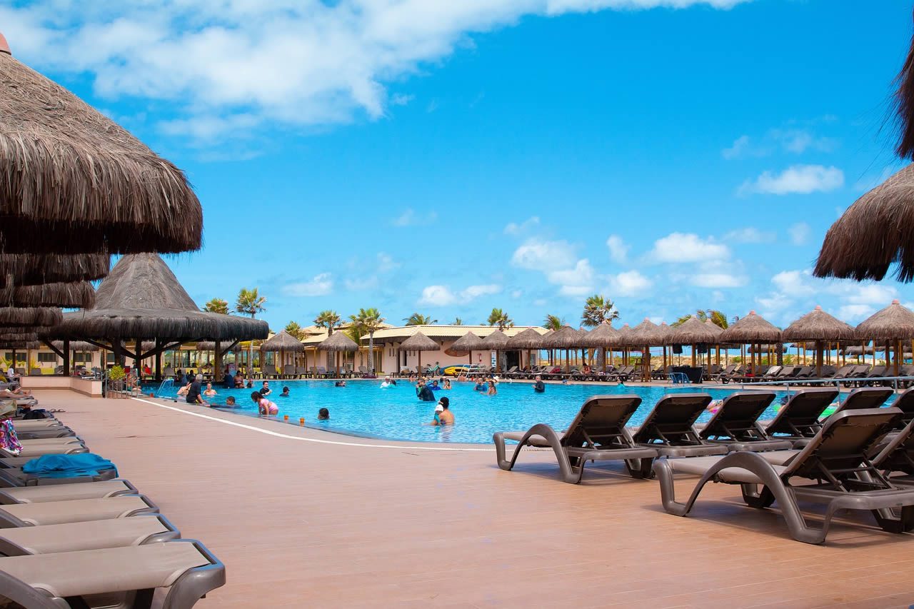 Holiday resort swimming pool with sun loungers, thatched parasols and people in the water under a bright blue sky