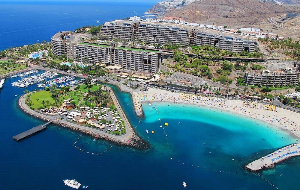 Aerial view of a large seaside resort complex with hotel buildings, marina, palm-lined pools and a sandy beach beside turquoise water and rocky coastline