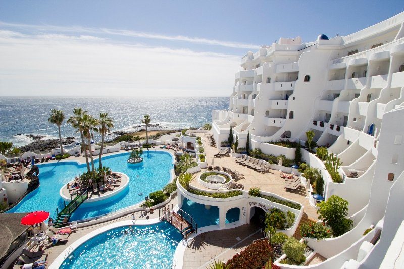 Seafront resort with white terraced apartments overlooking a turquoise swimming pool, sun loungers and palm trees by the ocean