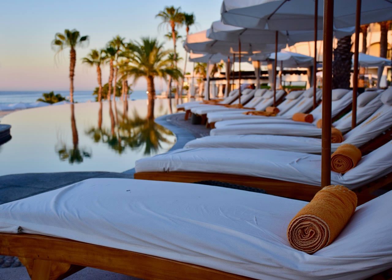 Sun loungers with rolled towels and white parasols beside an infinity pool with palm trees and sea view at sunset