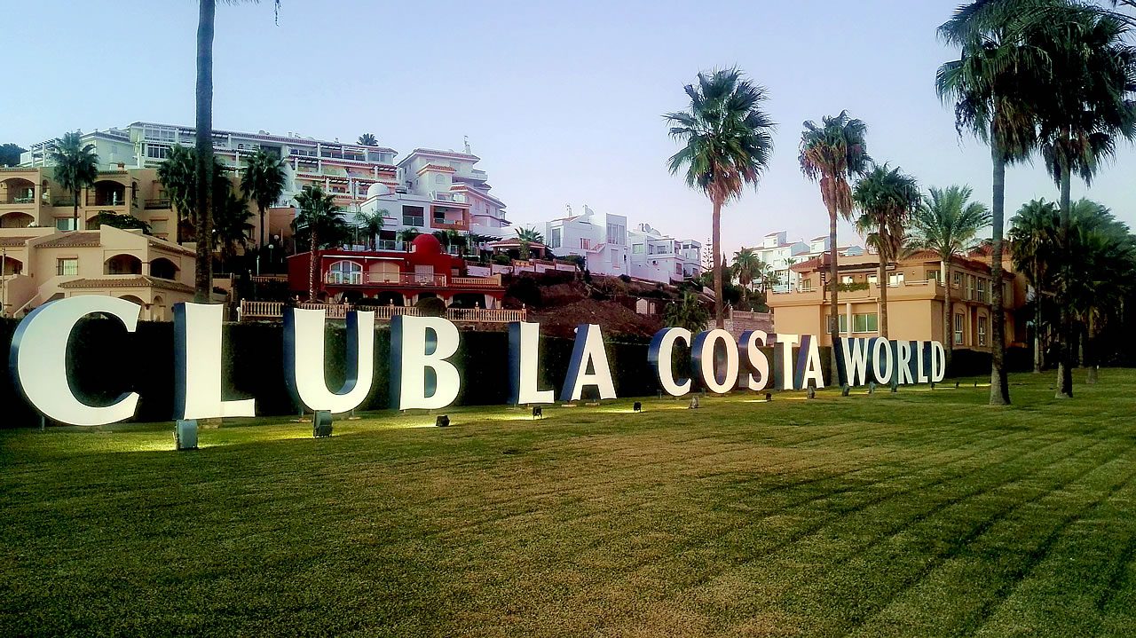 Large “CLUB LA COSTA WORLD” sign on a lawn with palm trees and resort apartment buildings in the background on the Costa del Sol, Spain
