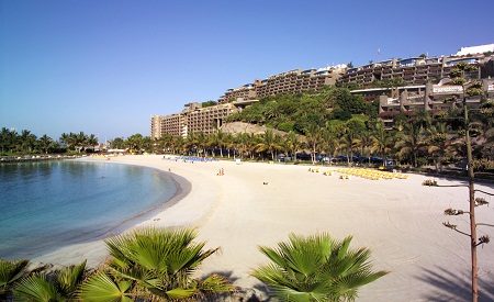 Wide view of a sandy beach with calm sea, palm trees and terraced resort buildings on a hillside under a clear blue sky