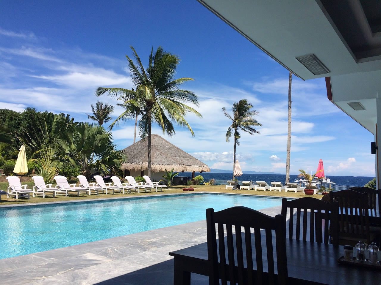 Infinity pool with sun loungers, palm trees and parasols overlooking the sea at a coastal resort, viewed from a shaded terrace dining area