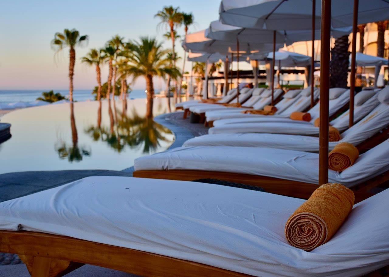 Sun loungers with rolled towels and umbrellas beside an infinity pool overlooking the sea and palm trees at a coastal resort