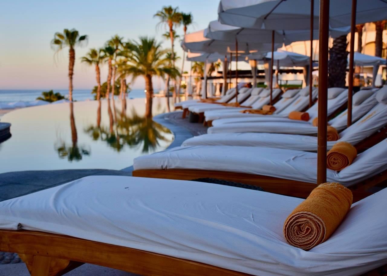 Sun loungers with rolled towels under parasols beside a beachfront infinity pool with palm trees at sunset