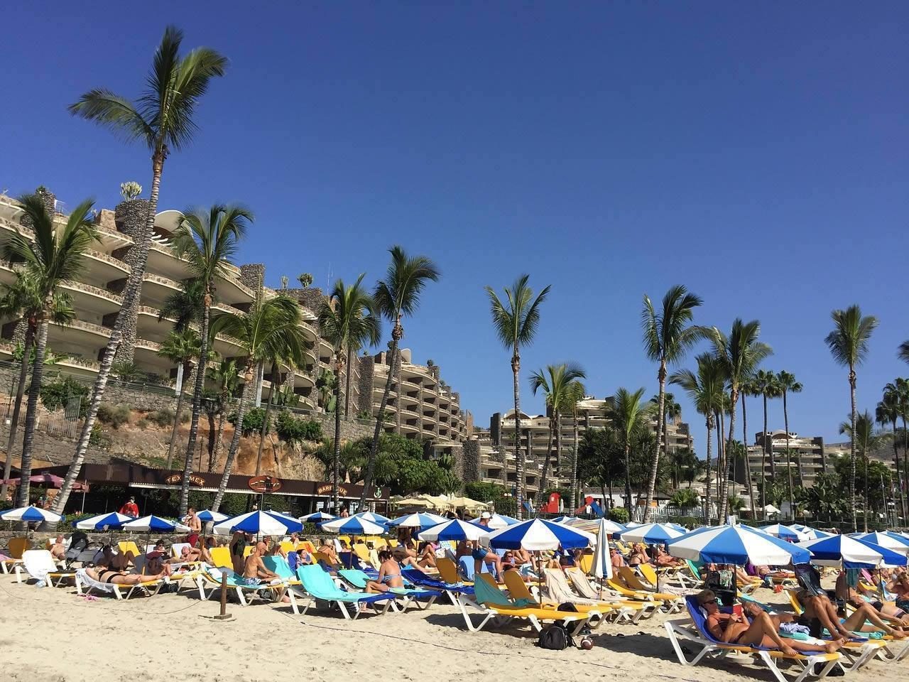 Sun loungers and blue-and-white parasols on a busy beach with palm trees and large resort buildings under a clear blue sky