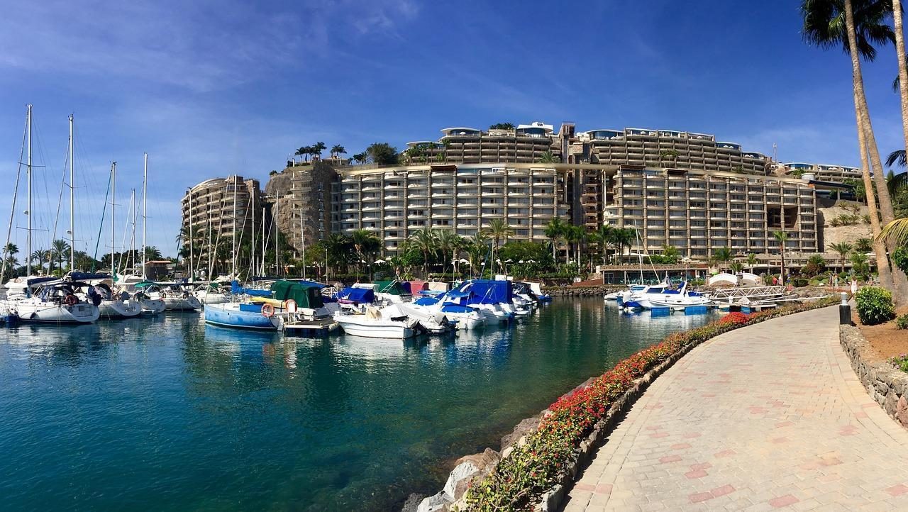 Marina with moored sailboats beside a waterfront promenade, with large resort apartment buildings and palm trees in the background under a clear blue sky