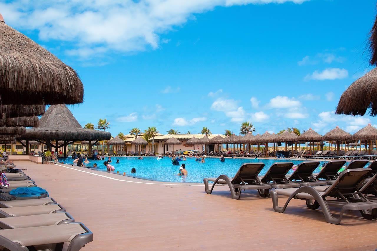 Sun loungers around a large resort swimming pool with thatched parasols and people swimming under a bright blue sky