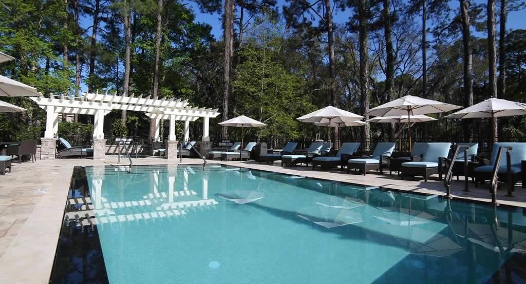 Outdoor resort swimming pool with sun loungers and parasols beside a white pergola, surrounded by tall trees