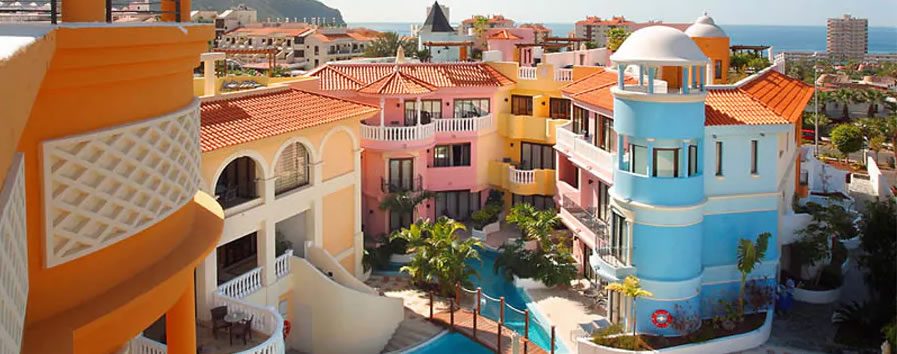 Colourful Mediterranean-style resort buildings with balconies and a turquoise pool, with the sea in the distance