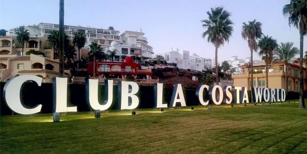 Large “CLUB LA COSTA WORLD” sign on a lawn with palm trees and resort buildings in the background